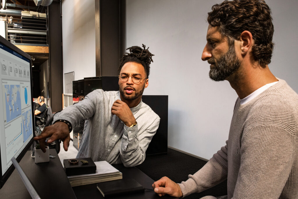 Man in sweater points at computer screen, showing something to bearded man in beige sweater.