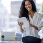 Young business woman using a digital tablet. She is casually dressed and smiling with a window behind her. She is sitting on a board room table with a laptop computer on it