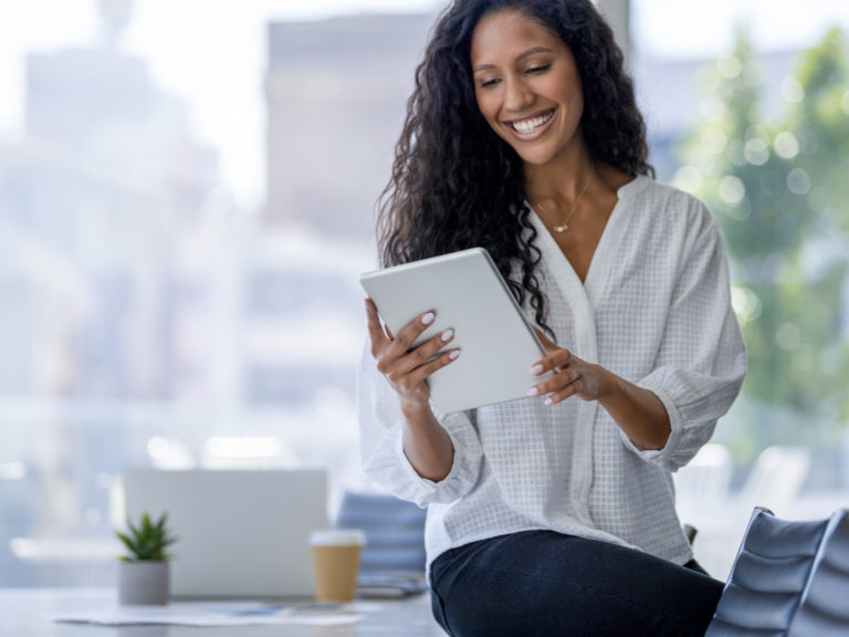 Young business woman using a digital tablet. She is casually dressed and smiling with a window behind her. She is sitting on a board room table with a laptop computer on it