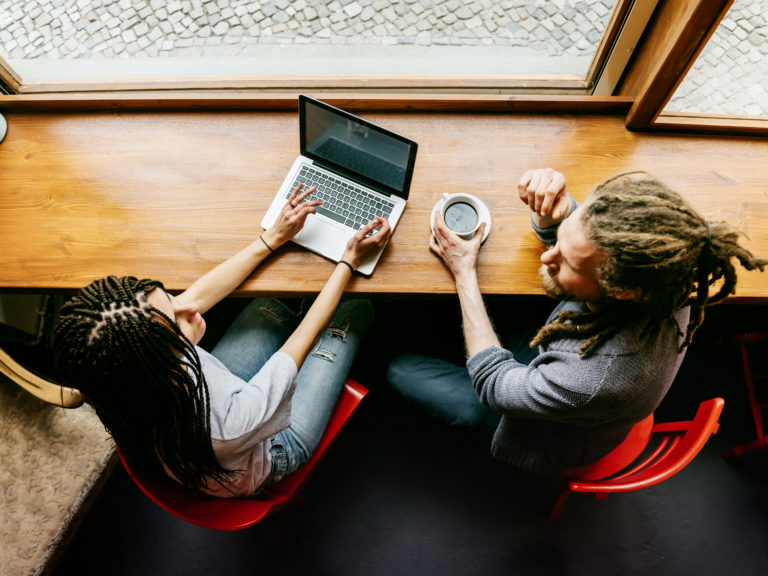A young man and a young woman are talking to each other in a bright café. Both of them have dark dreadlocks. She's working on a notebook while he is drinking some coffee.