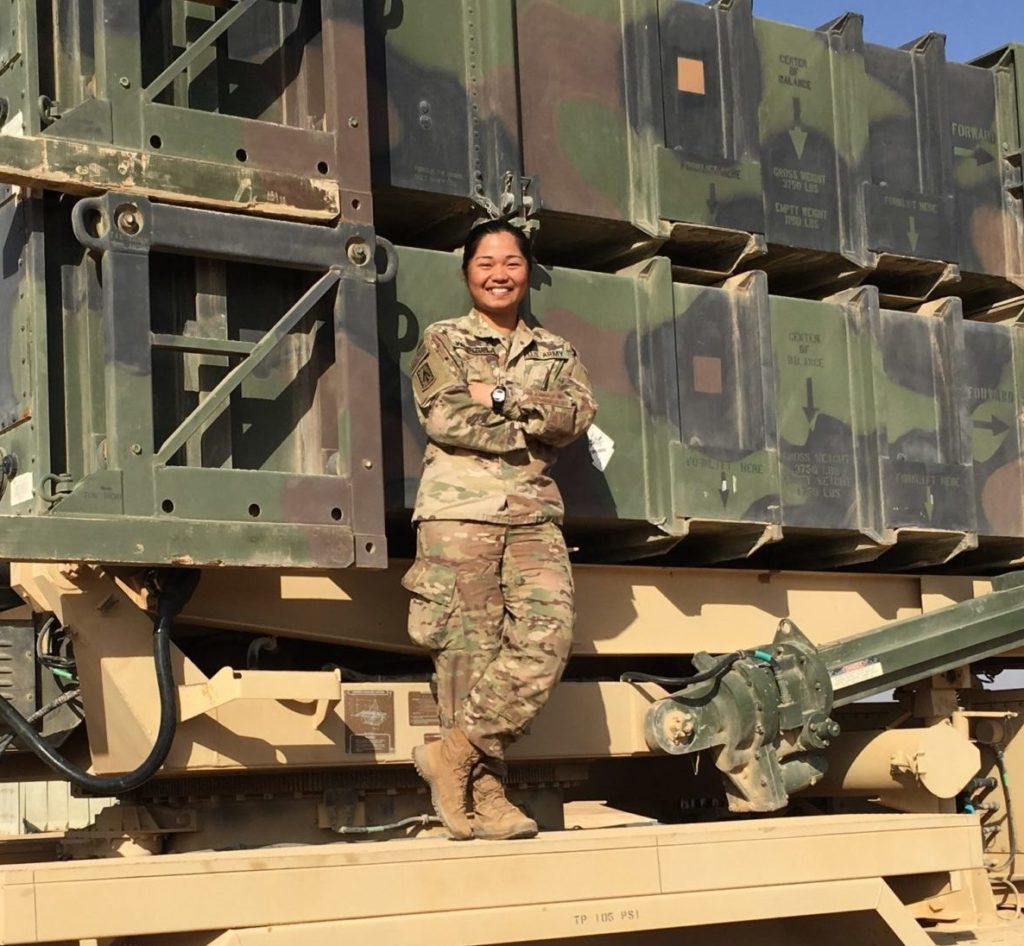 Female soldier stands in front of military transport truck in uniform, standing with arms crossed.